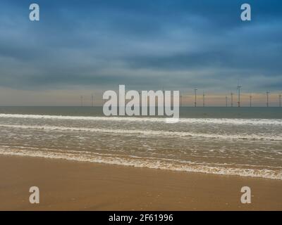 Le parc éolien proche de la rive à Redcar, avec des vagues se brisant sur les sables de plage en premier plan, le parc éolien à l'horizon et un ciel profondément coloré. Banque D'Images