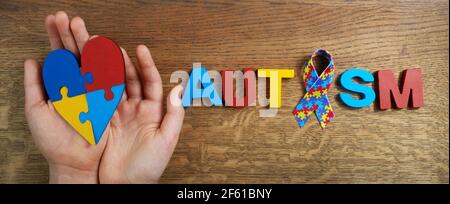 Les mains de garçon autiste tenant le coeur puzzle shapeed avec le mot autisme et le ruban d'awarennes sur fond de bois. Jour ou mois de sensibilisation à l'autisme. Banque D'Images