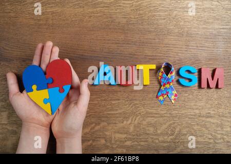 Les mains de garçon autiste tenant le coeur puzzle shapeed avec le mot autisme et le ruban d'awarennes sur fond de bois. Jour ou mois de sensibilisation à l'autisme. Banque D'Images