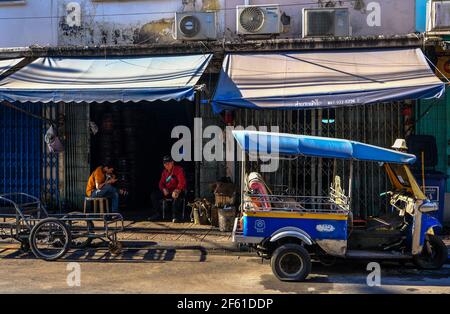 Deux hommes se détendent à l'extérieur de leur atelier derrière un tuk-tuk en stationnement, qui a besoin d'être réparé, à Talat Noi, Bangkok, Thaïlande Banque D'Images