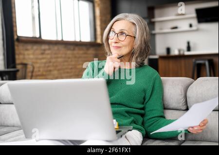 Femme séchée sur le canapé avec un ordinateur portable à la maison, elle tient du papier et prépare quelque chose, rêvant de passer des vacances Banque D'Images