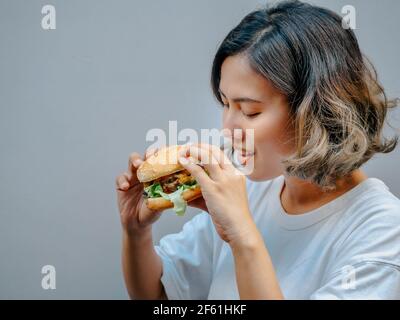 Délicieux hamburger maison frais. Belle femme asiatique heureuse cheveux courts portant un t-shirt blanc décontracté tenant et mangeant savoureux hamburger maison isola Banque D'Images