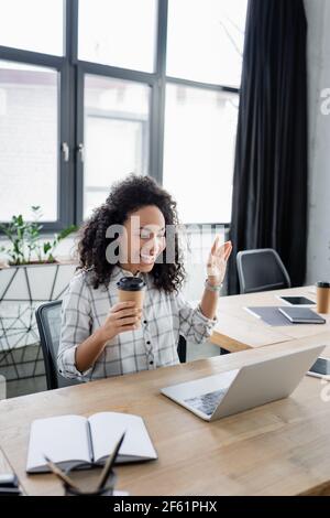 Femme d'affaires afro-américaine souriante avec boisson à emporter lors d'un appel vidéo sur ordinateur portable Banque D'Images