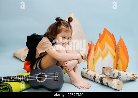 Petite fille confortable assise à côté d'un faux feu de camp avec des flammes de papier et des bûches de bouleau. Elle se serre les genoux et sourit. Sur fond bleu Banque D'Images