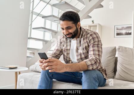 Un jeune indien heureux utilisant un smartphone assis sur un canapé à la maison. Banque D'Images
