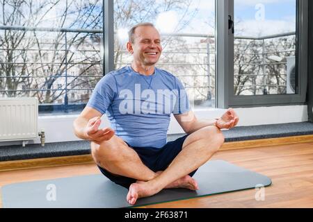 Homme d'âge moyen assis sur un tapis de sport dans un studio lumineux avec fenêtre panoramique, exercice de méditation de yoga à l'intérieur Banque D'Images