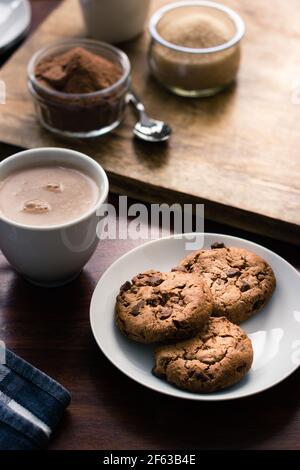 Pile de biscuits sur une assiette à côté d'une tasse de boire du chocolat avant des pots de sucre et de cacao en poudre Banque D'Images