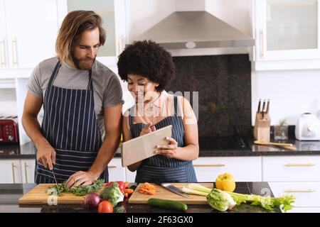 Joyeux couple varié dans la cuisine en utilisant une tablette et en préparant des aliments Banque D'Images