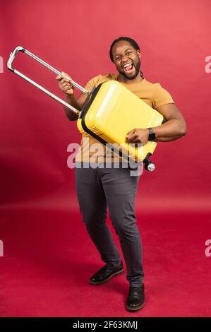 Joyeux gars afro-américain se tient avec une valise jaune, joue sur un sac comme un guitariste, se berner autour, un homme noir avec des bagages regardant un voyage, isolé sur rouge, pleine longueur Banque D'Images
