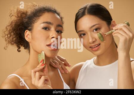 Soins de beauté. Jeunes femmes attrayantes regardant l'appareil photo, utilisant le rouleau de jade et le visage gua sha tout en posant ensemble isolé sur fond beige Banque D'Images