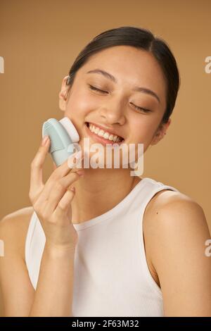 Belle jeune femme à l'air heureux tout en utilisant la brosse de nettoyage facial en silicone, posant isolé sur fond beige Banque D'Images