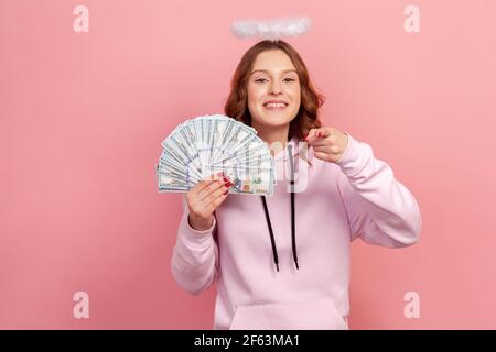 Portrait d'une adolescente heureuse à cheveux bouclés à capuche avec halo sur la tête tenant beaucoup de dollars et le doigt pointant sur l'appareil photo, Hé vous geste. Intérieur Banque D'Images