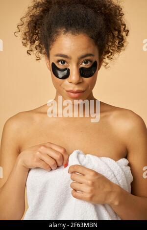 Portrait de la jeune femme enveloppée d'une serviette blanche après la douche pose avec application noire sous les cellules oculaires isolées au-dessus de beige arrière-plan Banque D'Images