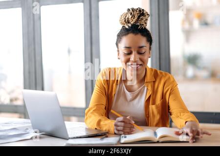 Formation en ligne. Joyeuse et intelligente Afro-américaine jeune femme élégante avec des dreadlocks, étudiante, étudier en ligne à la maison, regarder des leçons vidéo en ligne, utilise un ordinateur portable, prendre des notes, sourire Banque D'Images