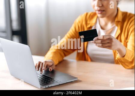 Achats et paiement en ligne. Femme afro-américaine défocuée dans des vêtements décontractés, assise à la table, payant pour des achats en ligne, tient une carte dans sa main, en saisissant les détails de la carte de crédit dans un ordinateur portable Banque D'Images