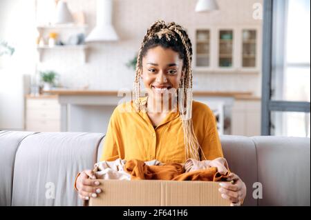 Joyeuse belle jeune africaine américaine avec des dreadlocks, assis à la maison dans le salon sur le canapé, tient boîte en carton avec des vêtements, regarde la caméra, souriant. Concept de livraison à domicile Banque D'Images