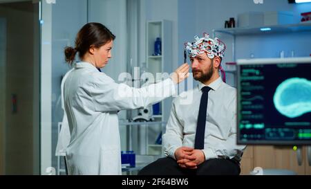 Homme assis sur une chaise neurologique avec un micro-casque à balayage d'ondes brainables tandis que le chercheur examine l'état de santé écrit sur le presse-papiers. Médecins travaillant dans un laboratoire d'étude du cerveau avec des moniteurs montrant la lecture EEG. Banque D'Images