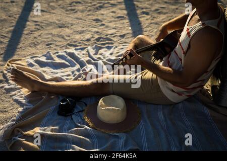 Partie basse de l'homme caucasien assis sur la plage à côté de guitare de mer Banque D'Images