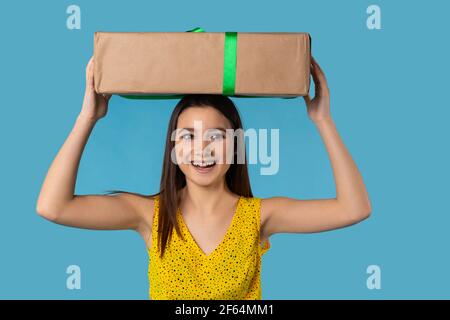 Image d'une dame excitée regardant côté debout sur fond bleu avec boîte de papier cadeau surprise avec ruban vert. Sourire une fille heureuse en été Banque D'Images