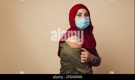 Portrait d'une femme musulmane montrant son bras avec un bandage après avoir été vaccinée. Femme islamique portant le hijab et le masque de visage regardant l'appareil photo sur marron Banque D'Images