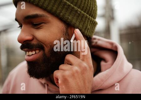 homme afro-américain souriant et barbu qui ajuste son écouteur tout en écoutant de la musique à l'extérieur Banque D'Images