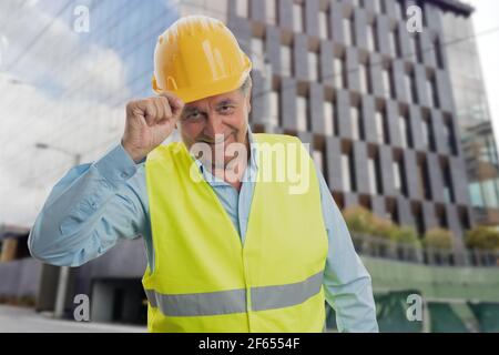 Ancien constructeur en travail gilet de tenue et casque jaune faire un geste de salutation respectueux toucher le casque avec la main souriante arrière-plan du bâtiment Banque D'Images