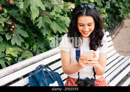 Une heureuse touriste qui utilise son téléphone à marteler assis sur un banc de parc. Accro à la technologie Banque D'Images