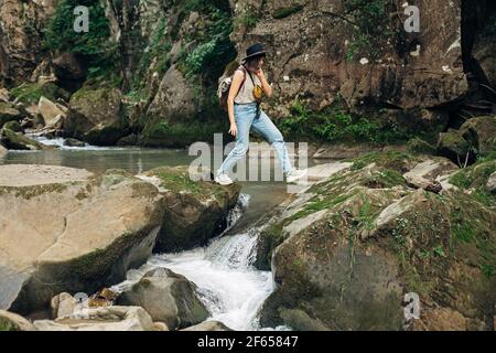 Femme élégante voyageur avec sac à dos marche sur les rochers de la rivière dans les montagnes. Jeune femme en tissu décontracté et chapeau sautant sur la rivière. Voyage et wa Banque D'Images