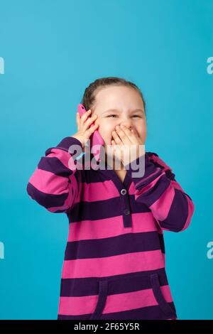 portrait d'une fille avec des picots riant aux larmes faisant appel au téléphone et couvrant sa bouche avec sa main de rire, isolé sur un fond bleu Banque D'Images