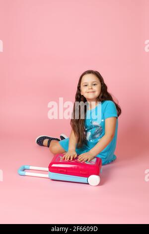 portrait complet d'une fille en robe bleue assise sur le sol avec ses mains sur une valise en plastique, isolée sur fond rose Banque D'Images