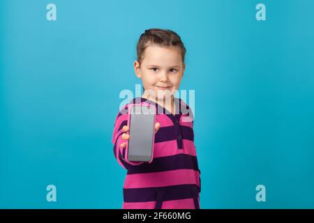 le portrait en gros plan d'une belle fille dans un sweat à capuche violet rayé montre l'écran du téléphone dans un cas rose, isolé sur un fond bleu Banque D'Images