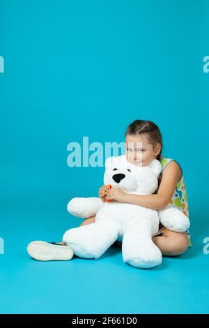 portrait d'une fille assise sur le sol, embrassant un ours en peluche blanc et jouant au dentiste tirant une dent avec des pinces isolées sur un fond bleu Banque D'Images