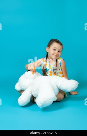 fille avec des queues de porc assis à pieds croisés sur le sol et jouant dentiste tirant une dent de bébé d'un ours en peluche blanc, isolé sur un fond bleu Banque D'Images