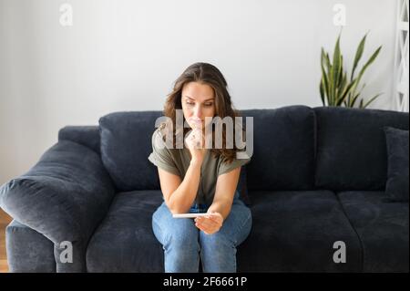 La jeune femme est assise sur le canapé à la maison avec un test de grossesse et attend le résultat, en attendant les manifestations de rayures Banque D'Images