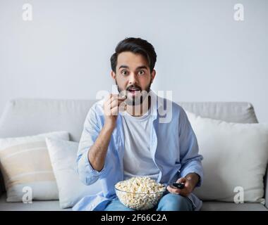 Jeune homme arabe enthousiaste regardant la télévision et mangeant du pop-corn à accueil Banque D'Images