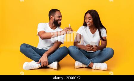 Couple noir utilisant des téléphones mobiles, assis au sol au studio Banque D'Images