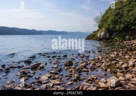 Rochers au bord du Loch Ness, en Écosse, par une journée ensoleillée et claire, avec les montagnes visibles sur la rive opposée Banque D'Images