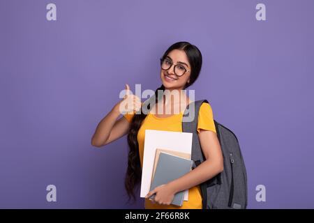 Joyeux étudiant indien portant des lunettes tenant des carnets montrant les pouces vers le haut Banque D'Images