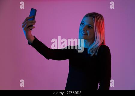 jeune femme blonde éclairée par des lumières néon prend un selfie avec son téléphone portable Banque D'Images