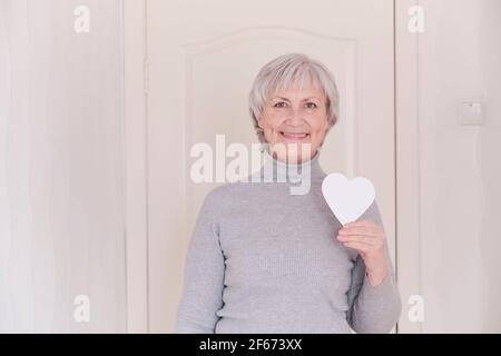 Un portrait d'une femme caucasienne âgée souriante avec de courts cheveux gris tenant dans votre main un petit coeur blanc, maquette. Banque D'Images