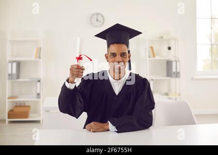 Joyeux étudiant en robe de remise des diplômes noire et chapeau assis à bureau et montrant son diplôme Banque D'Images