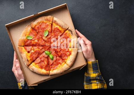 Mains de sexe masculin tenant la pizza pepperoni dans une boîte en carton sur fond de béton noir, vue de dessus. Service de livraison de pizzas. Copier l'espace pour le texte ou la conception Banque D'Images