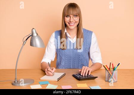 Photo portrait de l'assistant dans les lunettes écriture liste dans le copybook souriant au bureau, isolé sur fond beige pastel Banque D'Images