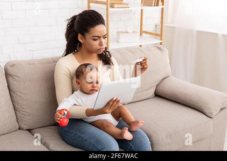 Une maman africaine frustrée avec un bébé malade utilisant une tablette à la maison Banque D'Images