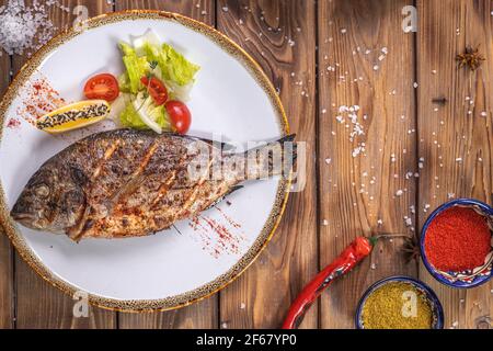 Poisson grillé sur une assiette de légumes, poivron, gros sel sur fond de bois brun. Service de restaurant. Banque D'Images