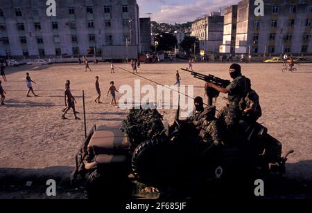 Les jeunes jouent au football dans un quartier appauvri de Rio de Janeiro, tandis que des militaires recherchent des trafiquants de drogue. Occupation par l'armée des zones défavorisées dans le cadre de la politique gouvernementale de sécurité publique en vue de la coupe du monde de la FIFA 2014 au Brésil. Banque D'Images