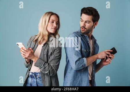 Jeune homme curieux et femme qui peeking dans leurs smartphones isolés sur fond bleu Banque D'Images