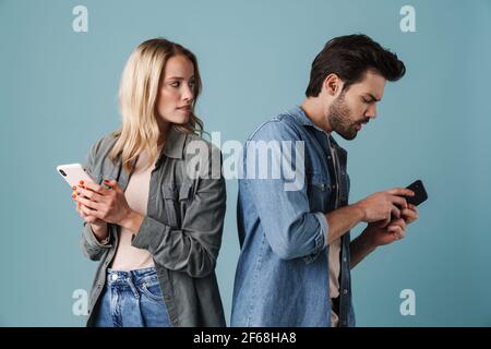 Jeune homme curieux et femme qui peeking dans leurs smartphones isolés sur fond bleu Banque D'Images