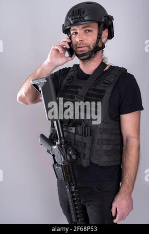 Un homme équipé pour Airsoft avec un casque et une veste de protection parle au téléphone en se tenant debout avec son arme exposée. Les communications sont importantes Banque D'Images