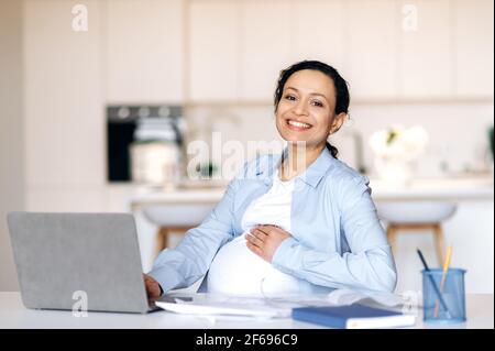 Bonne mignonne de course mixte femme enceinte adulte, femme d'affaires, designer ou directeur travaillant à la maison avec un ordinateur portable, assis au travail, regardant l'appareil photo, se portant le ventre, souriant amical Banque D'Images